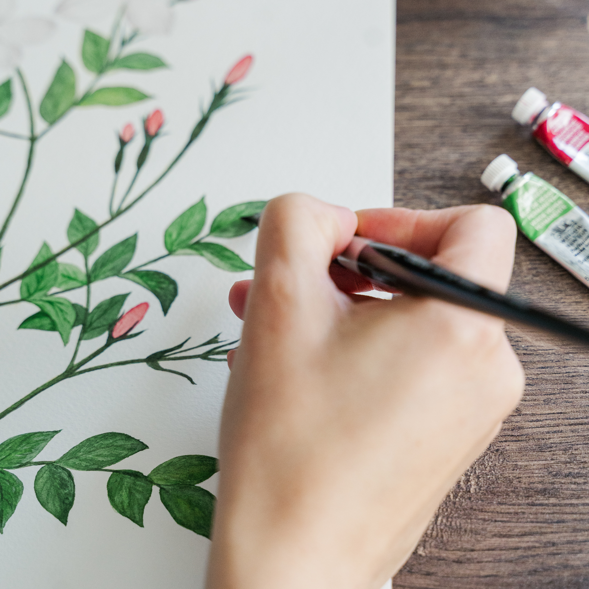 Hand painting a floral design on paper with paint tubes on a wooden surface.
